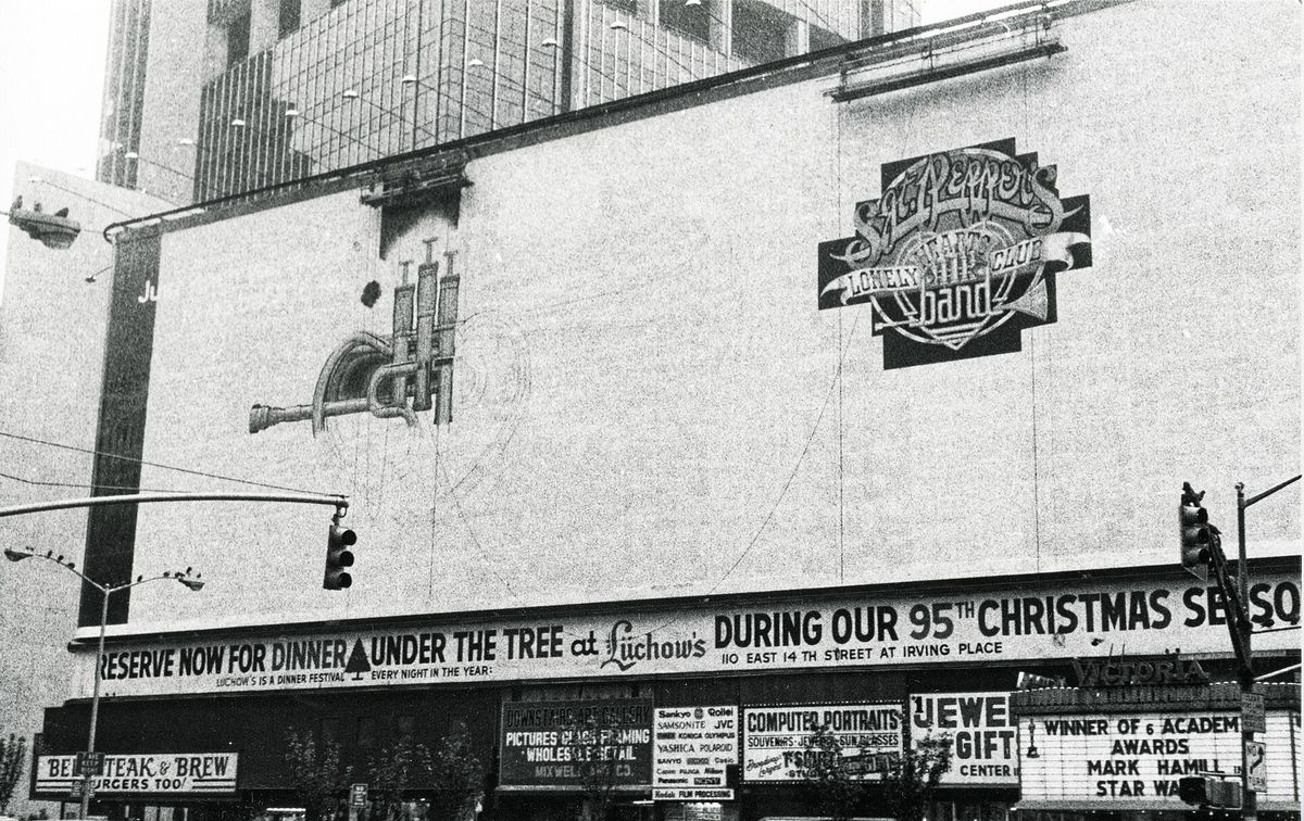 Black and white photo of a huge painted billboard for the Sgt. Pepper's Lonely Hearts Club Band film above a New York theater marquee, with street signs below