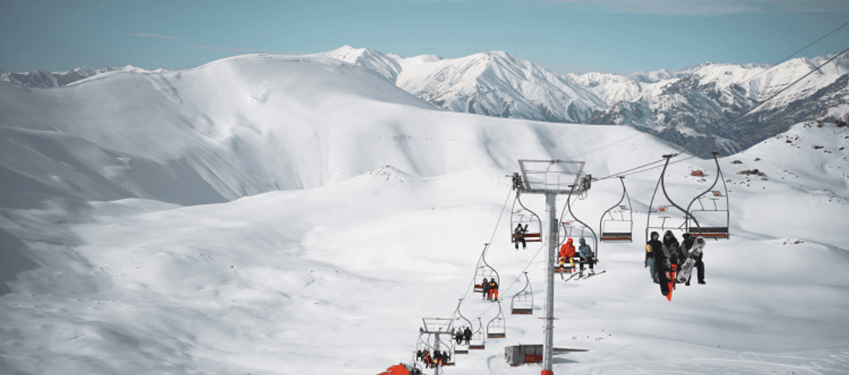Skiers on a lift with snowy mountain backdrop.