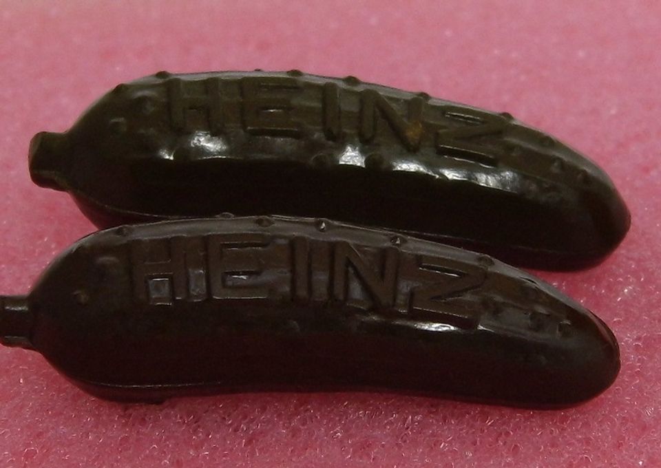 Close-up of two small dark green plastic pickle-shaped pins, each embossed with the word HEINZ, lying on a pink foam surface