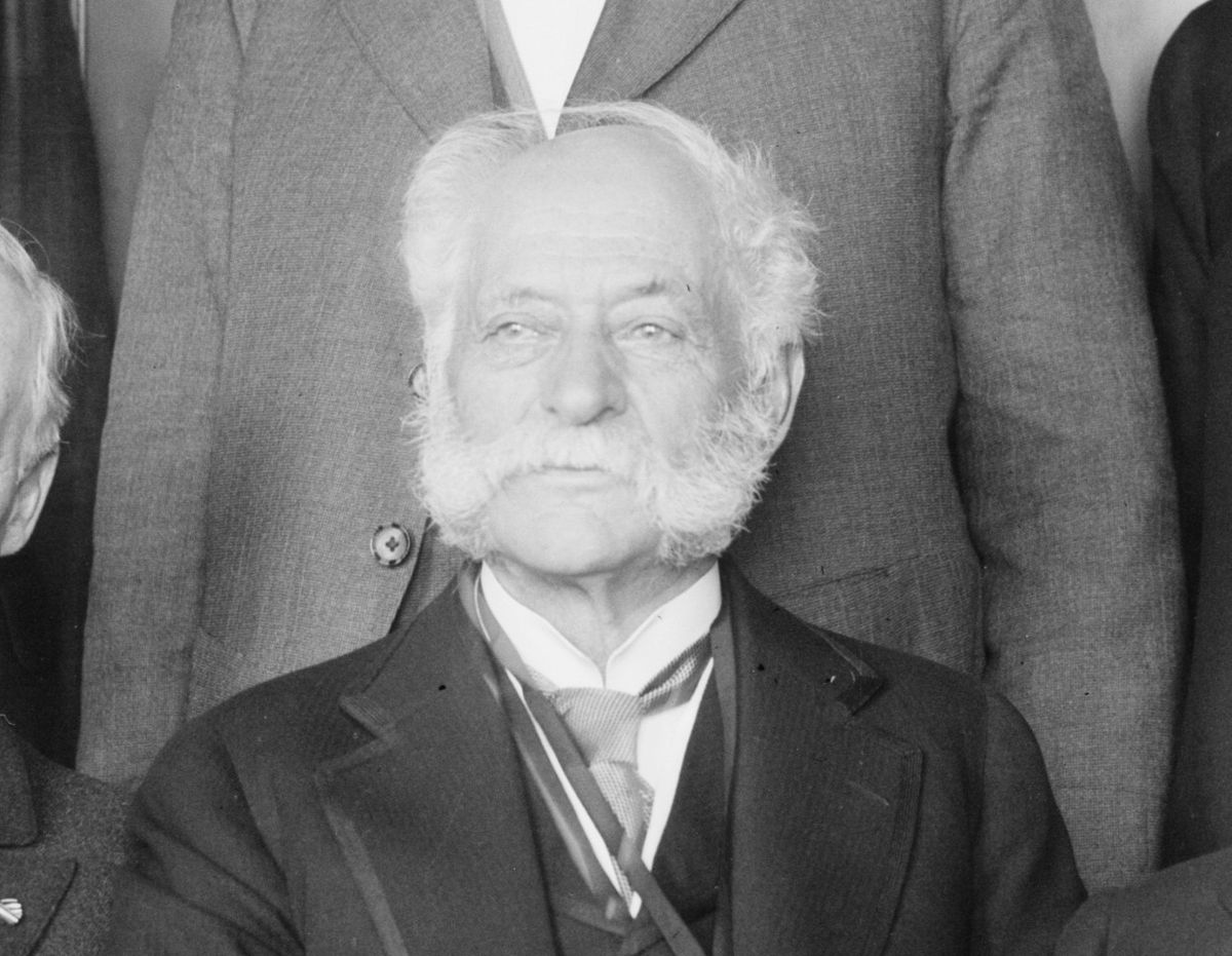 Black and white photograph of an elderly Henry J. Heinz with white hair and large white side whiskers, wearing a dark suit with a ribbon around his neck, seated among other men