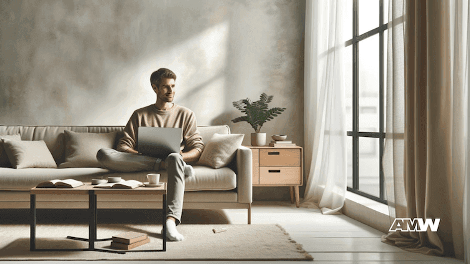 A man with a laptop sitting on a sofa in a quiet grey living room beside a tall curtained window