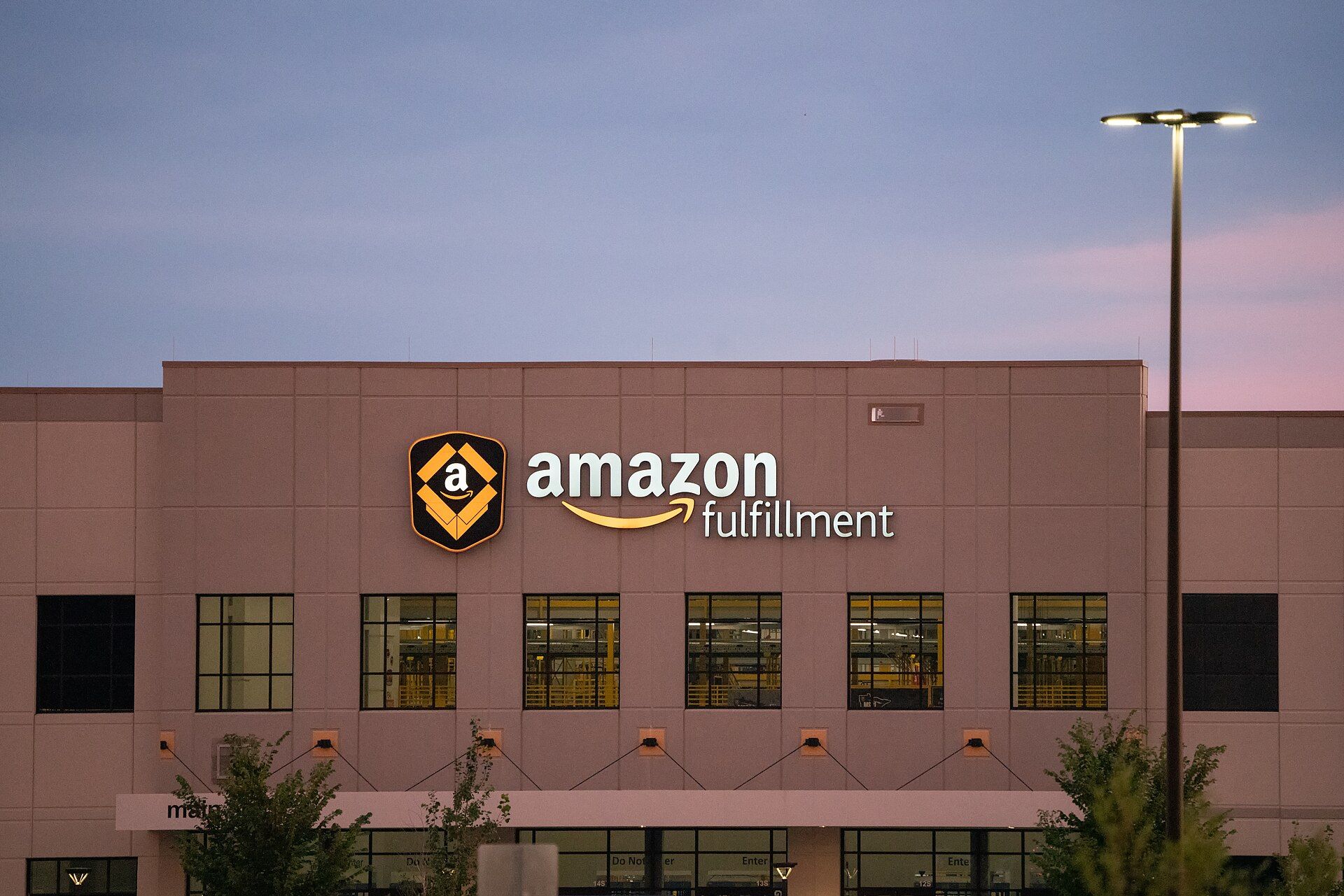 Exterior of a tan warehouse at dusk with an illuminated amazon fulfillment sign and logo above a row of lit windows, with a tall street light at right