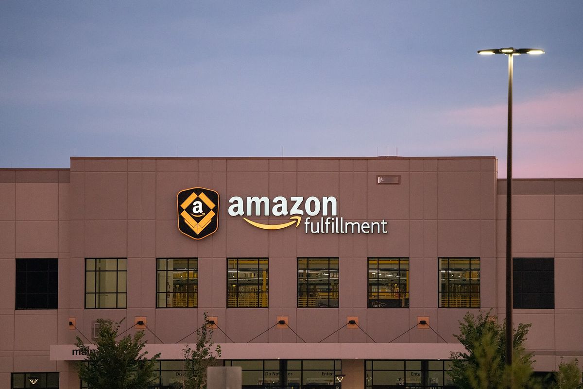 Exterior of a tan warehouse at dusk with an illuminated amazon fulfillment sign and logo above a row of lit windows, with a tall street light at right