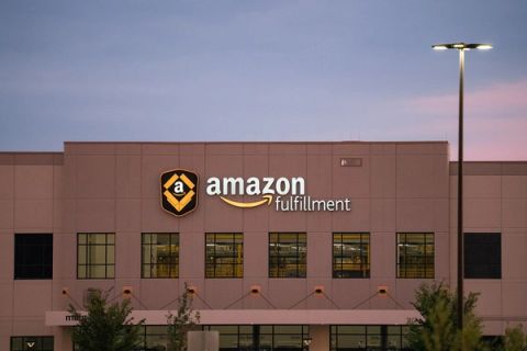 Exterior of a tan warehouse at dusk with an illuminated amazon fulfillment sign and logo above a row of lit windows, with a tall street light at right