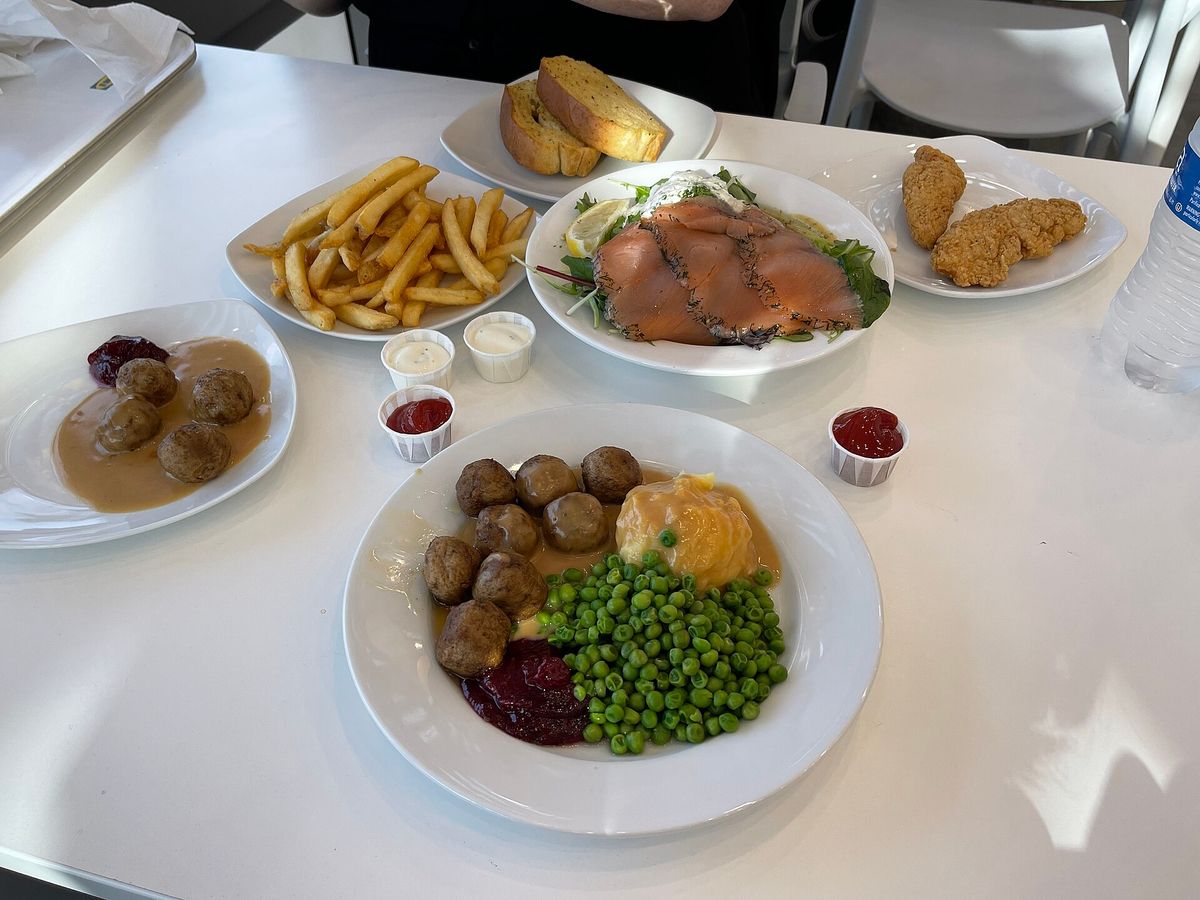 A white table in an IKEA restaurant with plates of meatballs with mashed potatoes, peas and lingonberry jam, a plate of smoked salmon on greens, french fries, fried chicken strips, garlic bread and small cups of ketchup and sauce