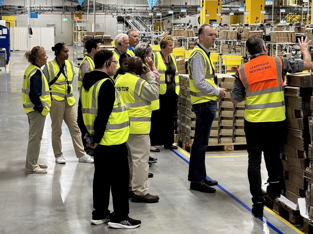 A group of visitors in yellow safety vests listening to a guide in an orange vest beside stacks of flattened cardboard boxes inside a large warehouse with conveyor lines overhead