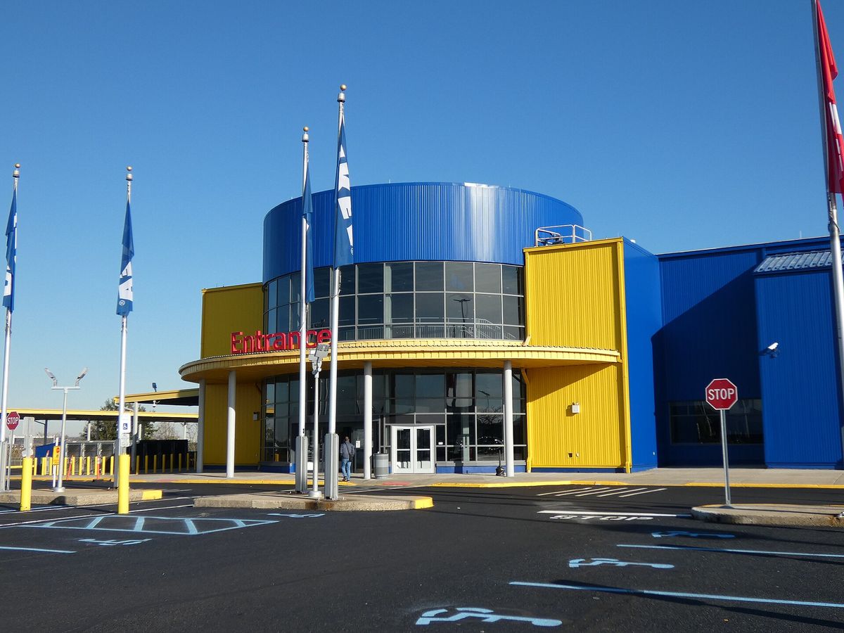 The entrance of a blue and yellow IKEA store with a round blue tower, a red Entrance sign, flagpoles and an empty parking lot under a clear sky