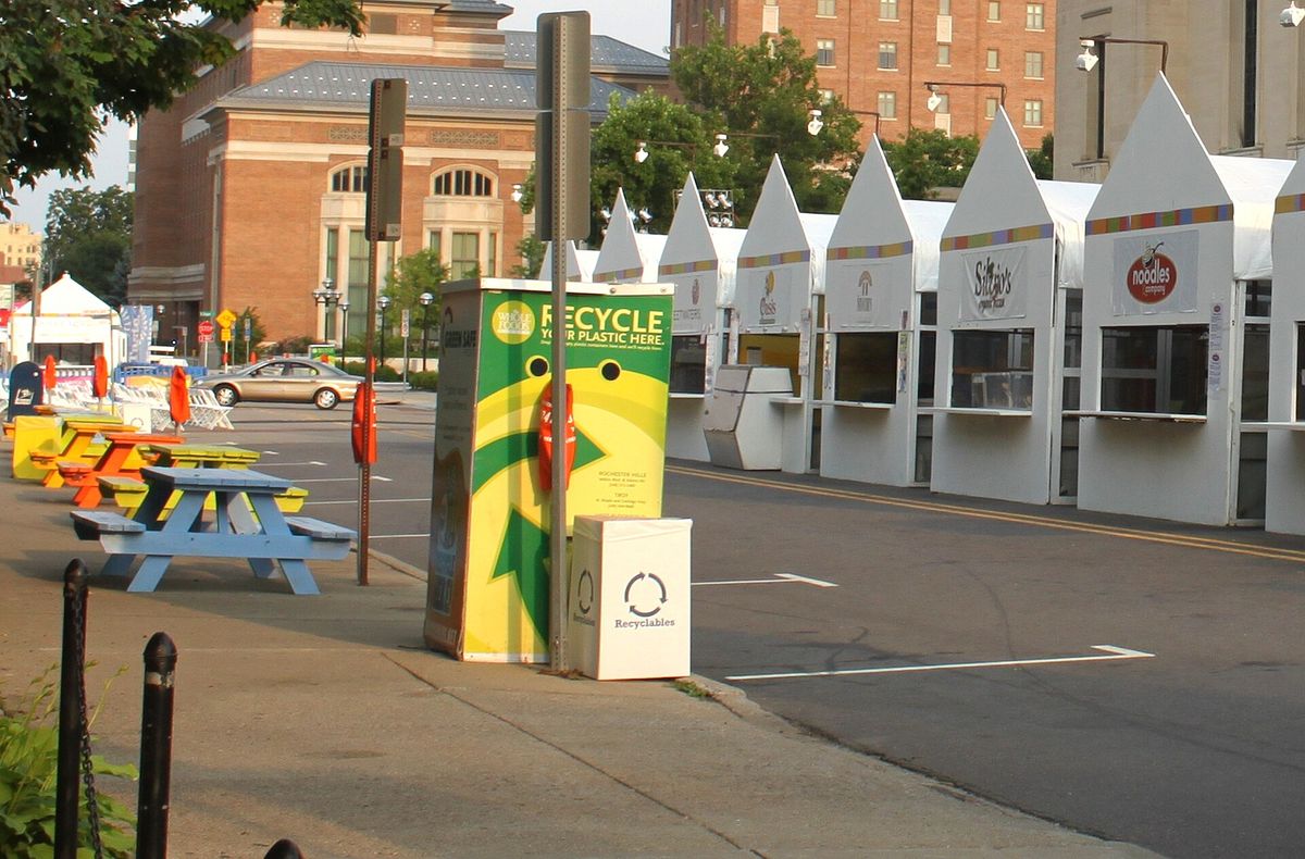 A green and yellow recycling bin labeled Recycle your plastic here with the Whole Foods Market logo, on a sidewalk beside picnic tables and white festival booths
