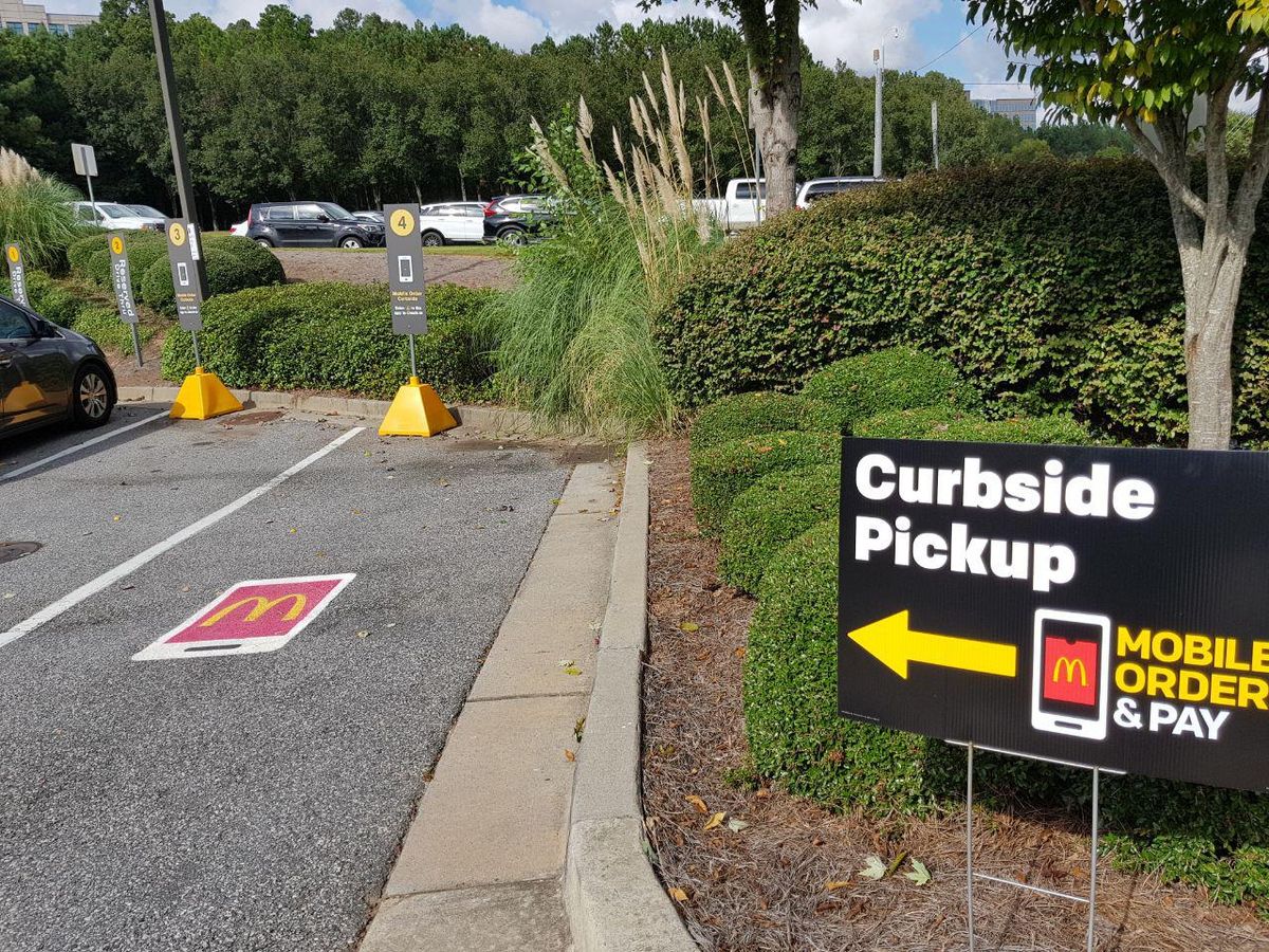 A black sign reading Curbside Pickup with a yellow arrow and a phone icon labeled Mobile Order and Pay, beside a parking lot with numbered pickup bays and a McDonald's logo painted on the asphalt