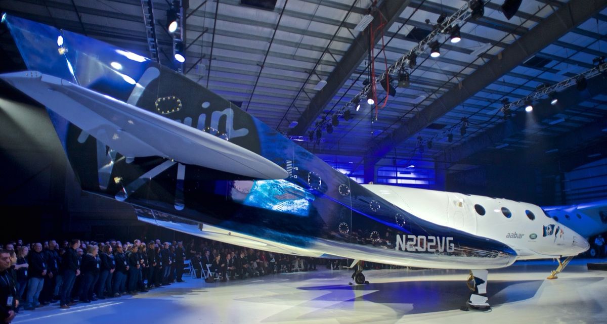 White and blue Virgin Galactic spaceship with registration N202VG on display in a hangar in front of a large crowd