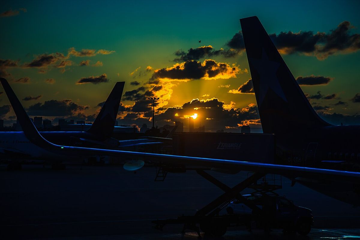 Silhouetted airliner tails and a catering truck on the apron at Miami International Airport at sunset, with the city skyline behind