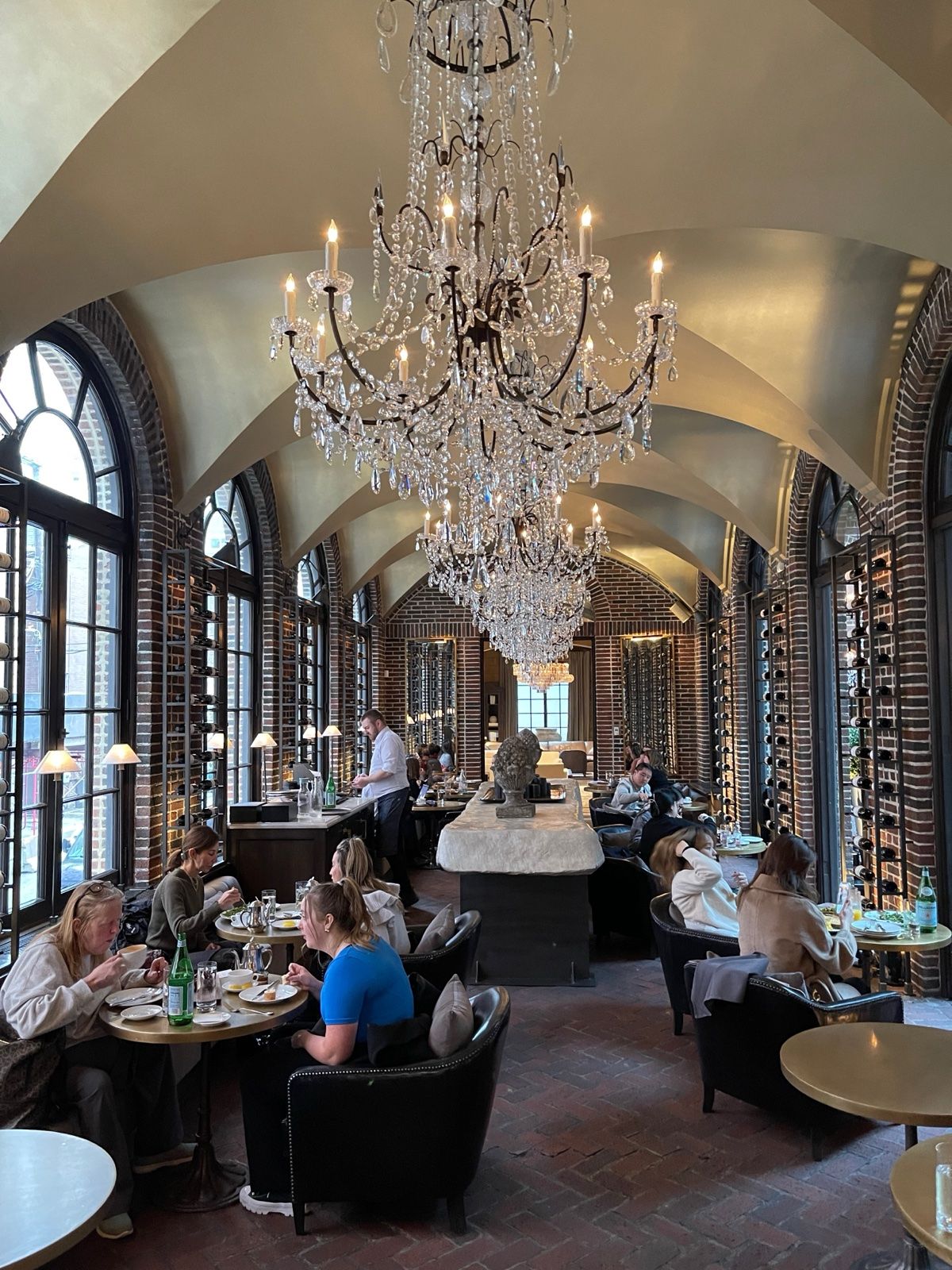 Long vaulted dining room with brick walls, arched windows, crystal chandeliers, wine racks and diners seated at small round tables