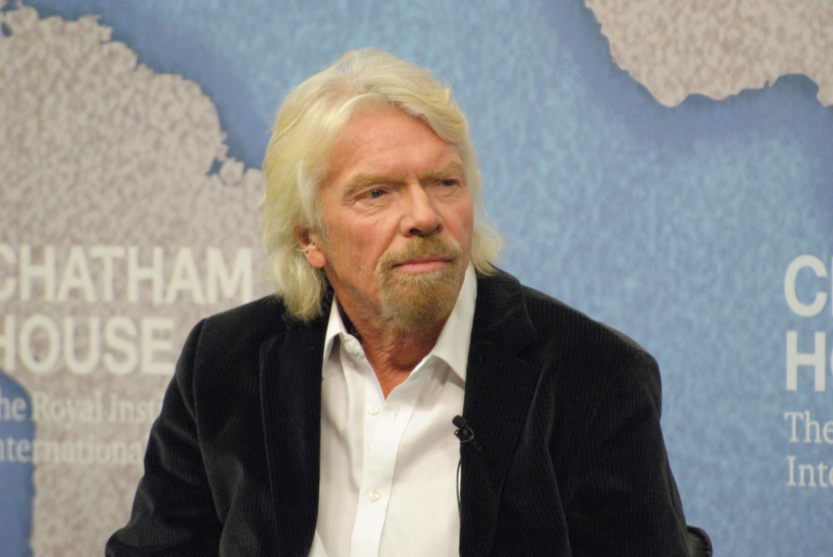 Richard Branson with white hair and a goatee, wearing a dark velvet jacket and white shirt, seated in front of a blue Chatham House backdrop