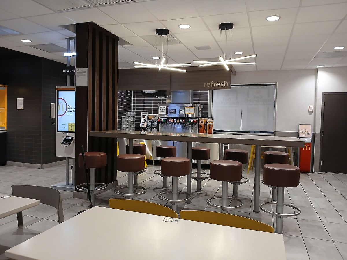 Inside a McDonald's dining room, a self-order kiosk on the left shows a screen reading Earn delicious rewards, next to a high counter with brown stools, a drink station and pendant lights