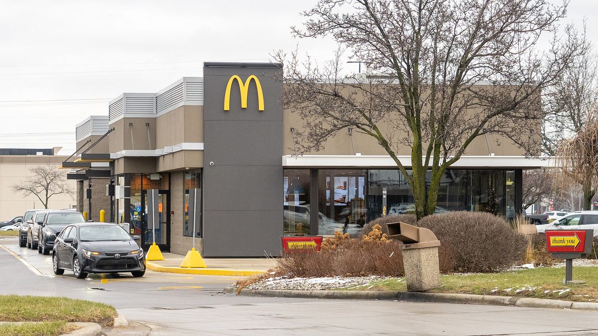 Cars lined up in the drive-thru lane of a McDonald's restaurant with a dark tower carrying the Golden Arches logo, bare trees and an overcast sky, Macomb County, Michigan