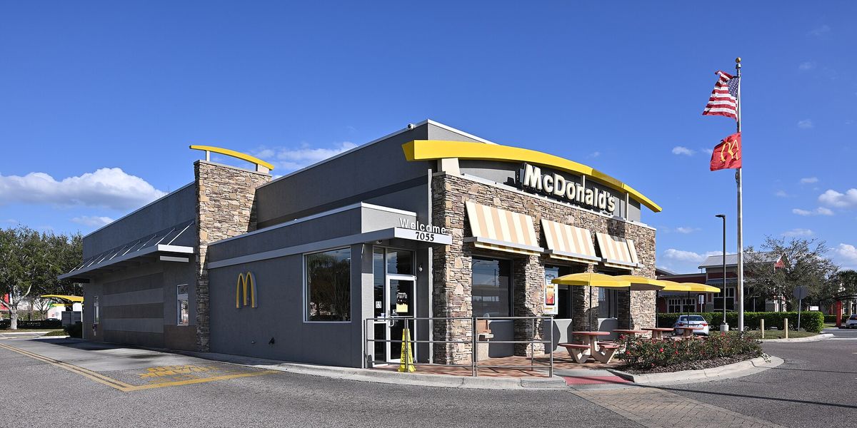 A single-storey McDonald's restaurant with a stone facade, yellow arch-shaped awnings over a patio and two flags on a pole, on a sunny day with a nearly empty parking lot