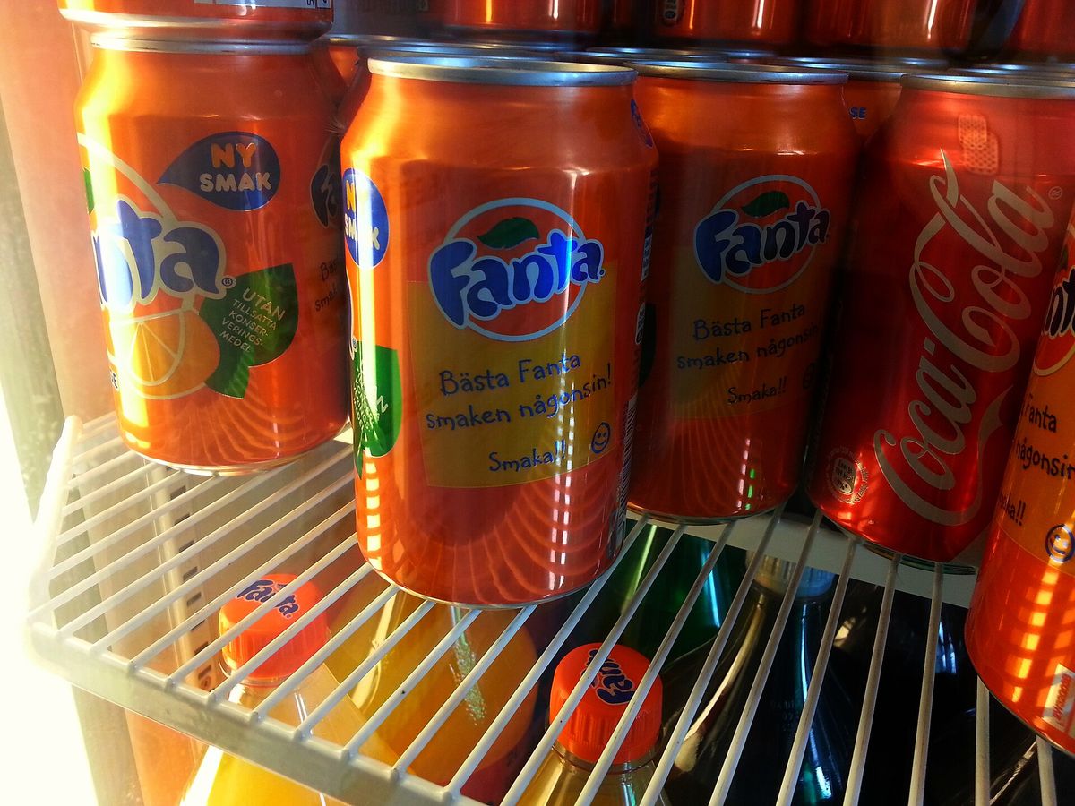 Orange Fanta cans with Swedish text stacked on a fridge shelf next to red Coca-Cola cans