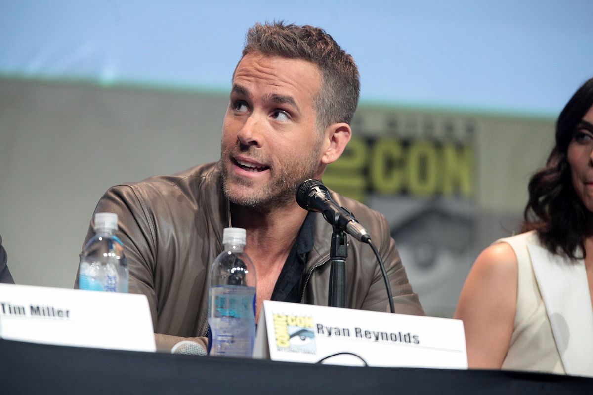 Ryan Reynolds, in a brown leather jacket, speaking into a microphone at a panel table with a name card in front of him at San Diego Comic-Con
