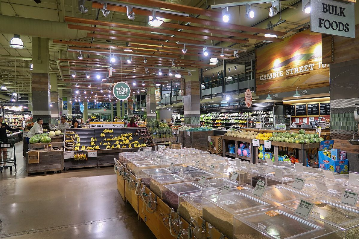 Rows of clear bulk food bins in front of banana and melon displays, with hanging signs reading Whole Trade and Bulk Foods, inside a large store
