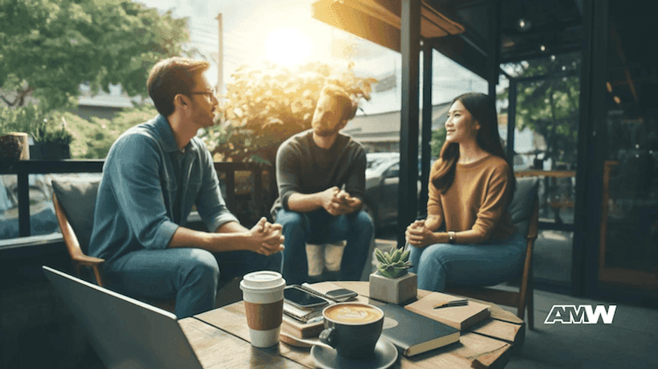 Three people sitting and talking around a café table outdoors, with a laptop, phones, notebooks and coffee cups in front of them