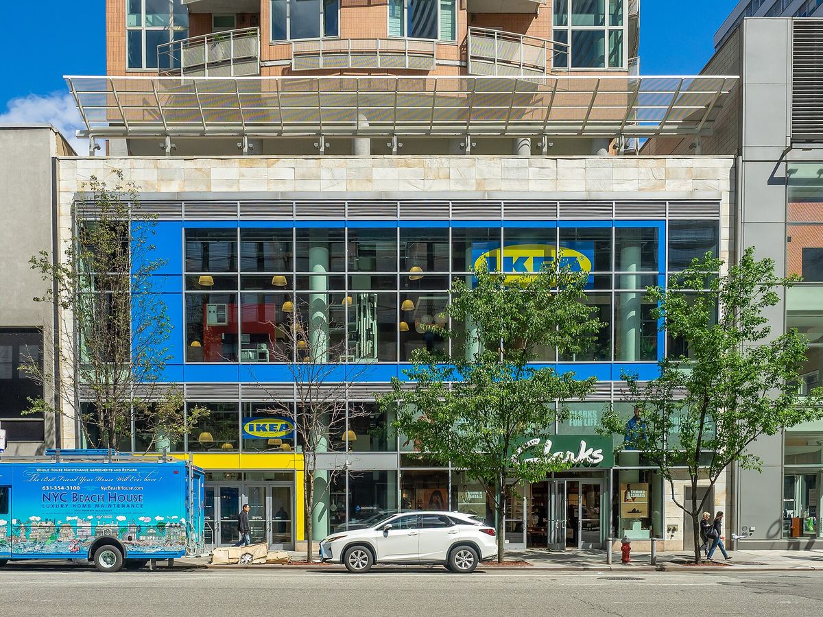 A city street front with a two-story glass window framed in blue and a yellow IKEA logo, part of a tall residential building, with pedestrians and a parked white SUV outside