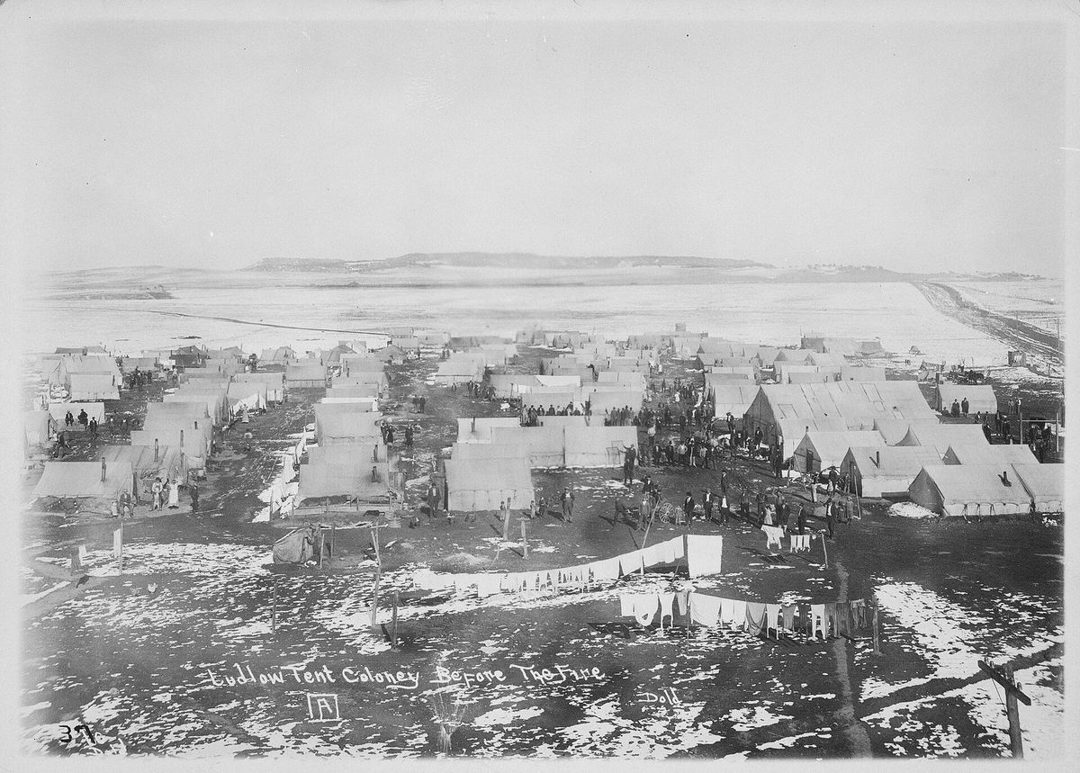 Wide black-and-white view over rows of white canvas tents on snowy ground, with laundry on lines in the foreground and open plains behind, captioned "Ludlow Tent Colony Before The Fire"