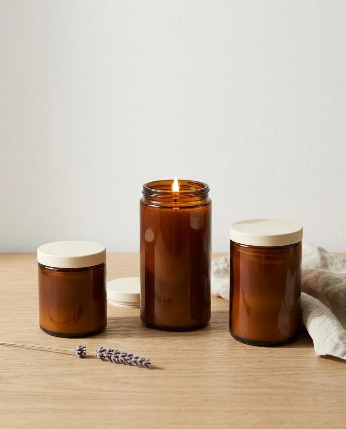 gemini-nano-banana-2.1 output: The photograph shows three amber glass candle jars of descending and ascending heights arranged on a light wooden surface against a plain off-white wall. The tall center candle is lit, while the left and right jars are closed with cream lids, accompanied by a lavender sprig and textured cloth.