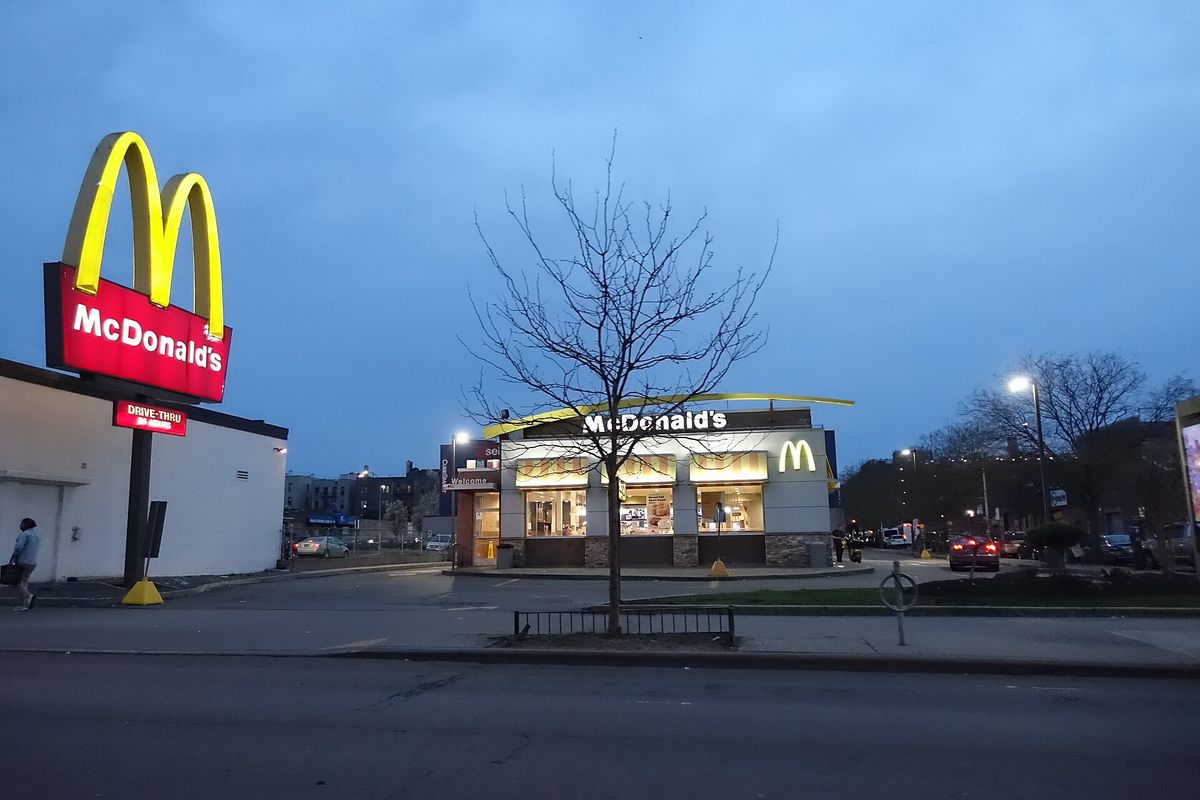 A tall McDonald's sign with the Golden Arches at dusk on a street corner, with a single-storey McDonald's restaurant lit up behind a bare tree across the parking lot