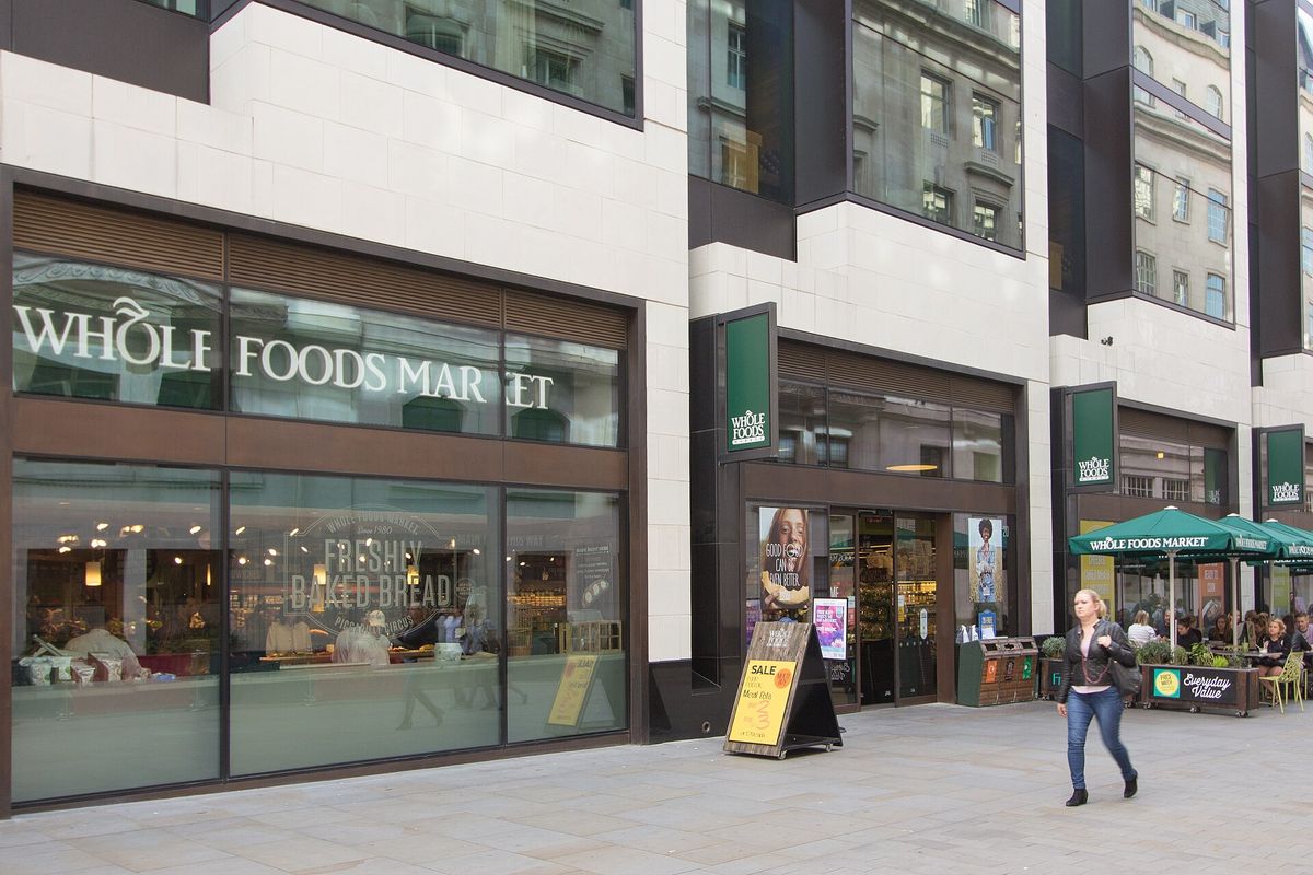 Street front of a Whole Foods Market in London, with large Whole Foods Market lettering over a bakery window, green hanging signs and green umbrellas over outdoor tables