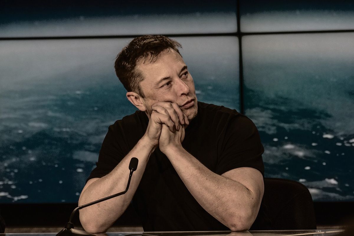 Elon Musk in a black T-shirt sitting at a table with his hands clasped under his chin, a microphone in front of him and an image of Earth's clouds on the screen behind