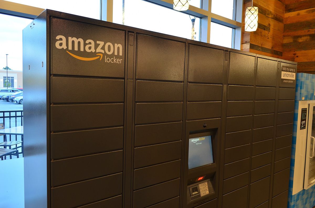A bank of dark grey Amazon Locker parcel compartments with a touchscreen kiosk, beside large windows inside a store