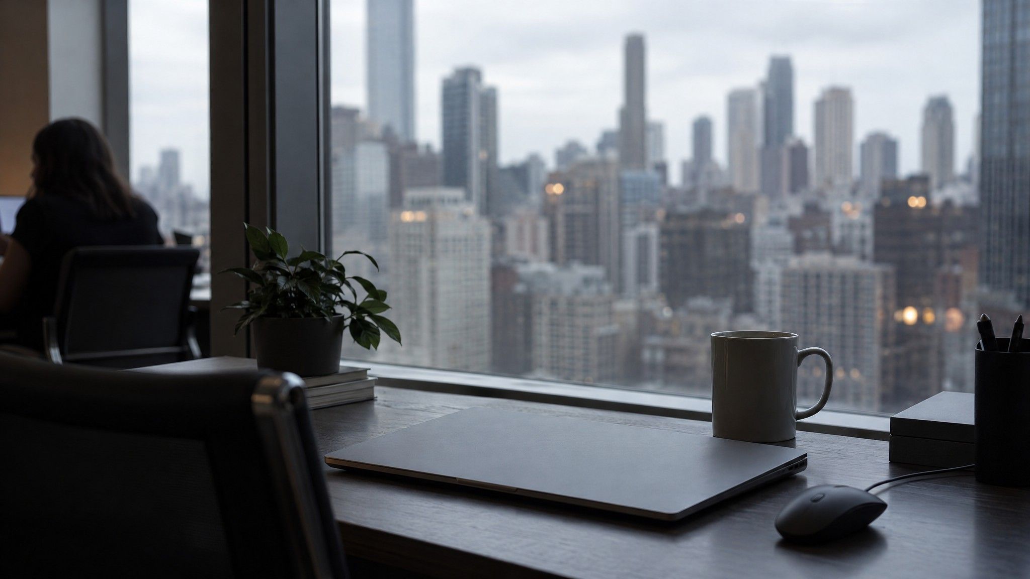 Closed laptop and mug on a dark desk with city windows behind