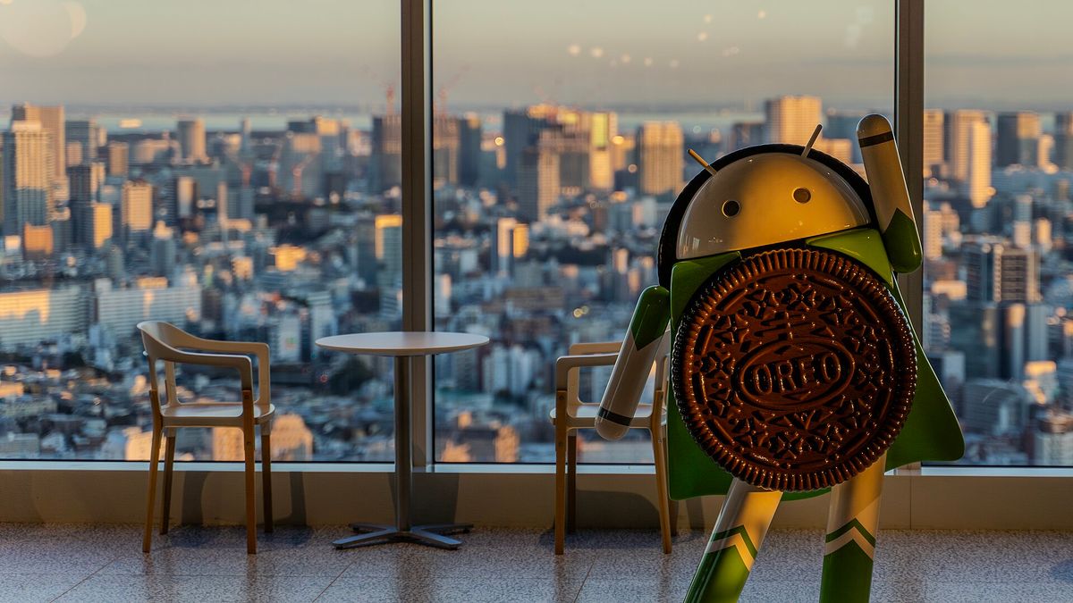 A green Android robot statue holding a large Oreo cookie in front of a window overlooking a city skyline