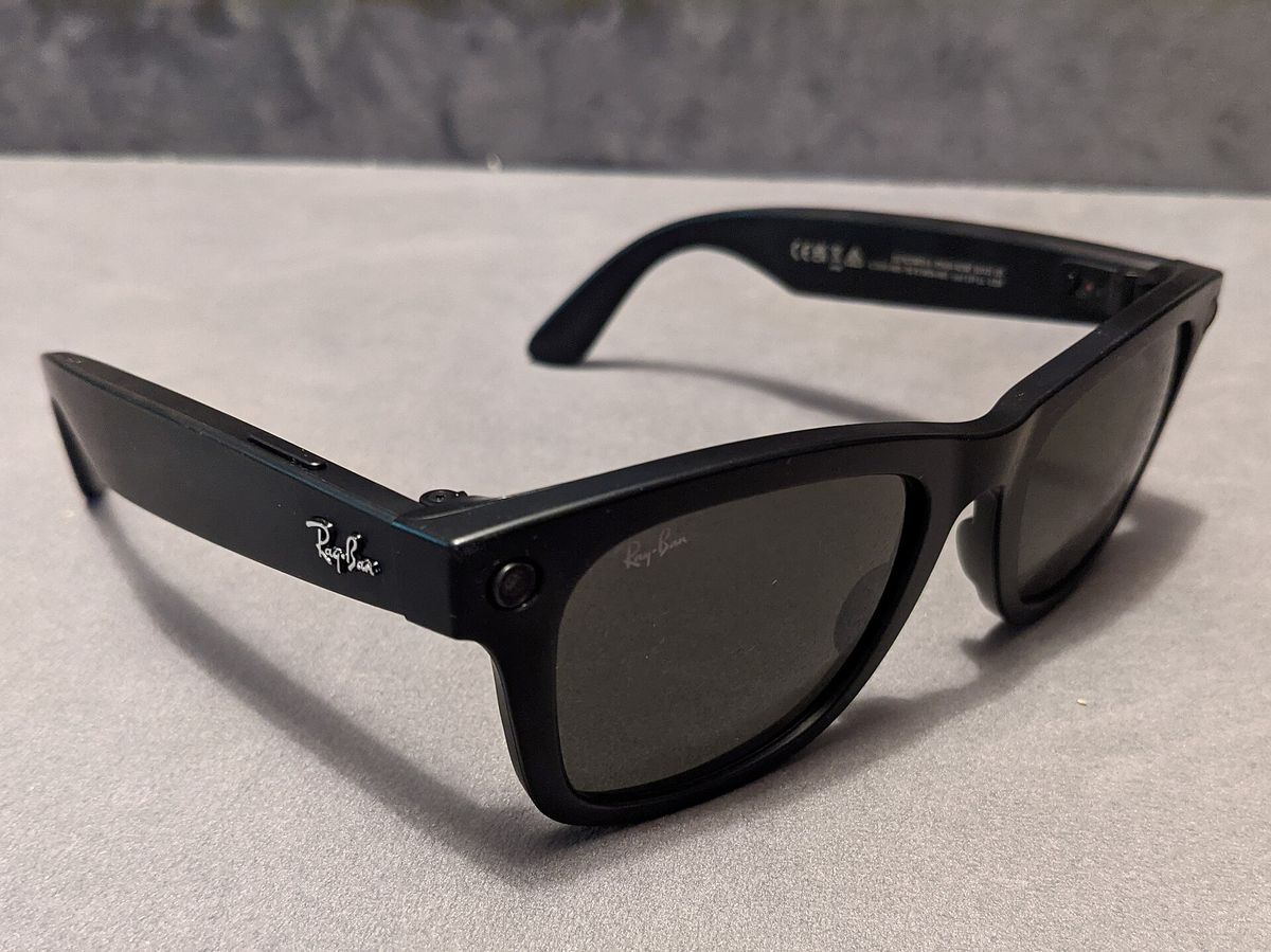 Black Ray-Ban Stories Wayfarer smart glasses resting on a grey table, with a camera lens at the corner of the frame