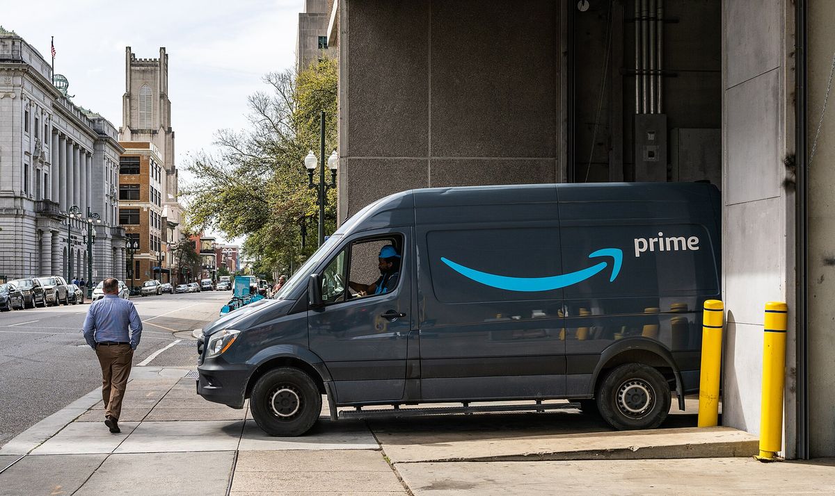 A dark gray delivery van with a blue smile logo and the word prime parked at a loading dock on a city street while a man walks past on the sidewalk