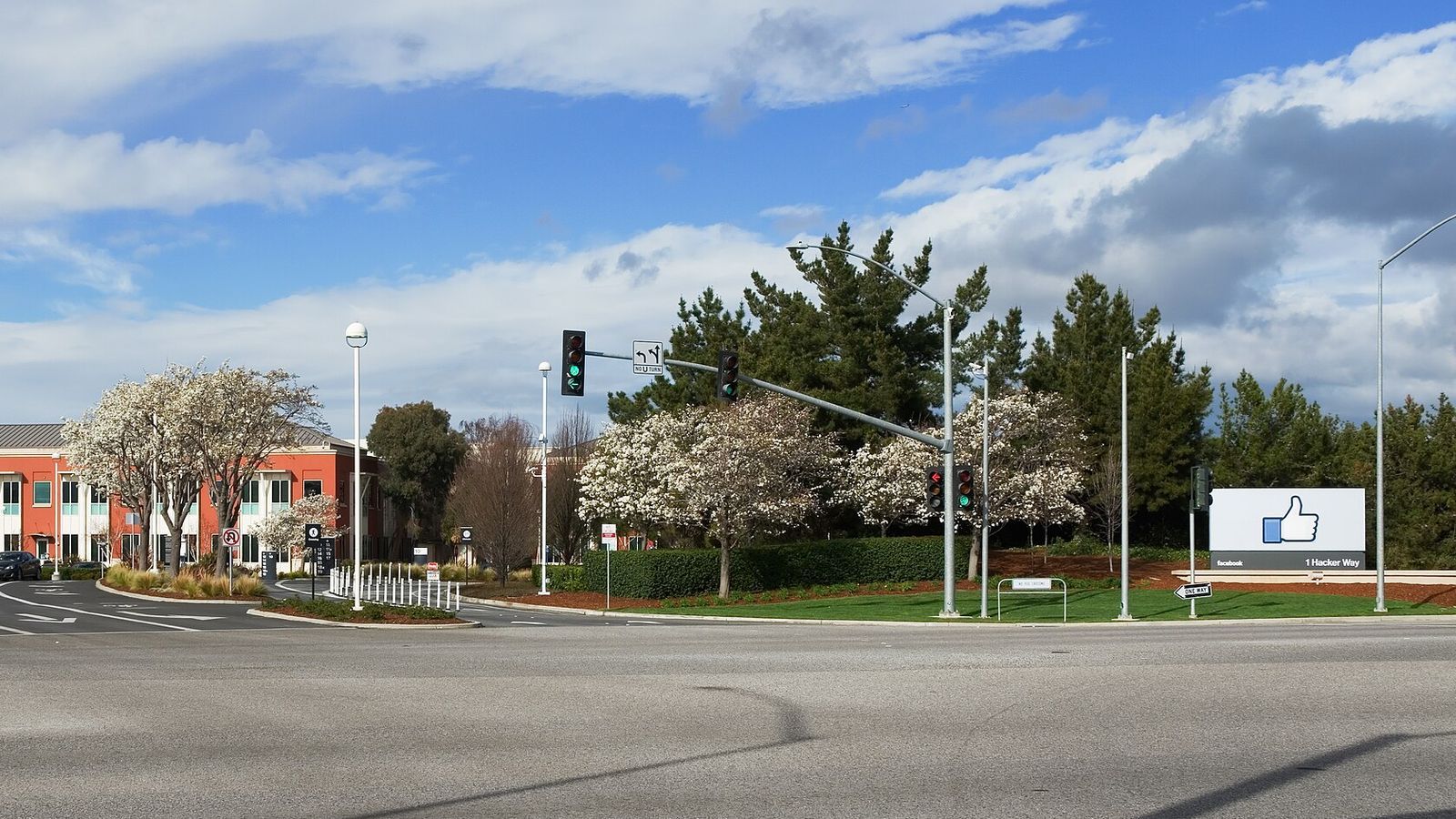 The entrance to Facebook's headquarters in Menlo Park, California, with a thumbs-up sign reading 1 Hacker Way, flowering trees and a red office building beside an intersection