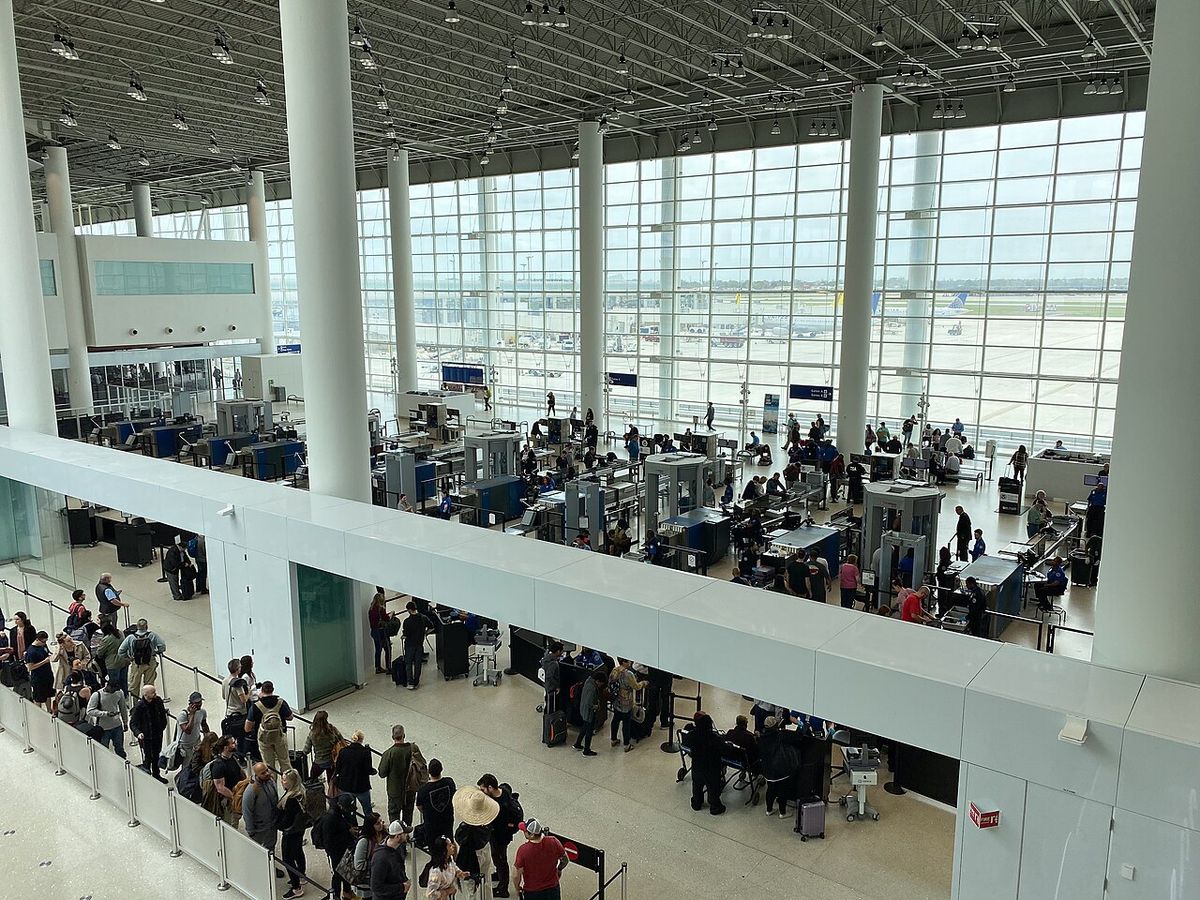 Overhead view of a busy public TSA security checkpoint with passengers queuing beside tall windows overlooking the airfield