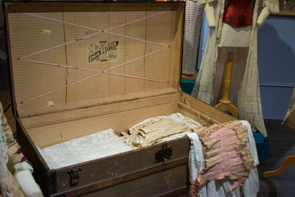 An open antique wooden trunk with a Louis Vuitton label on the striped paper lining of its lid, holding folded lace and linen, with dressed mannequins behind it