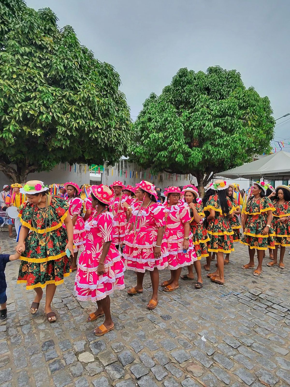 A group of women in bright pink and green floral dresses and matching hats walking along a cobbled street under two large green trees