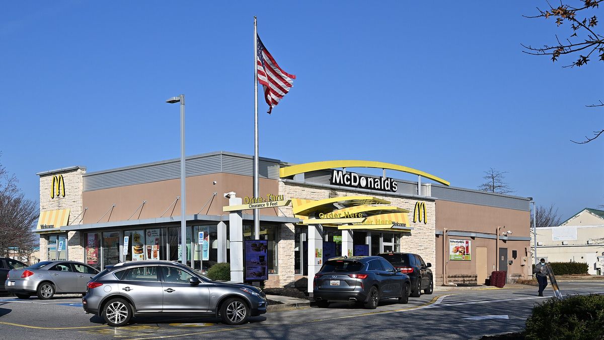 A stand-alone McDonald's restaurant with a yellow arch along its roofline and an American flag on a pole, with cars parked in front under a clear blue sky