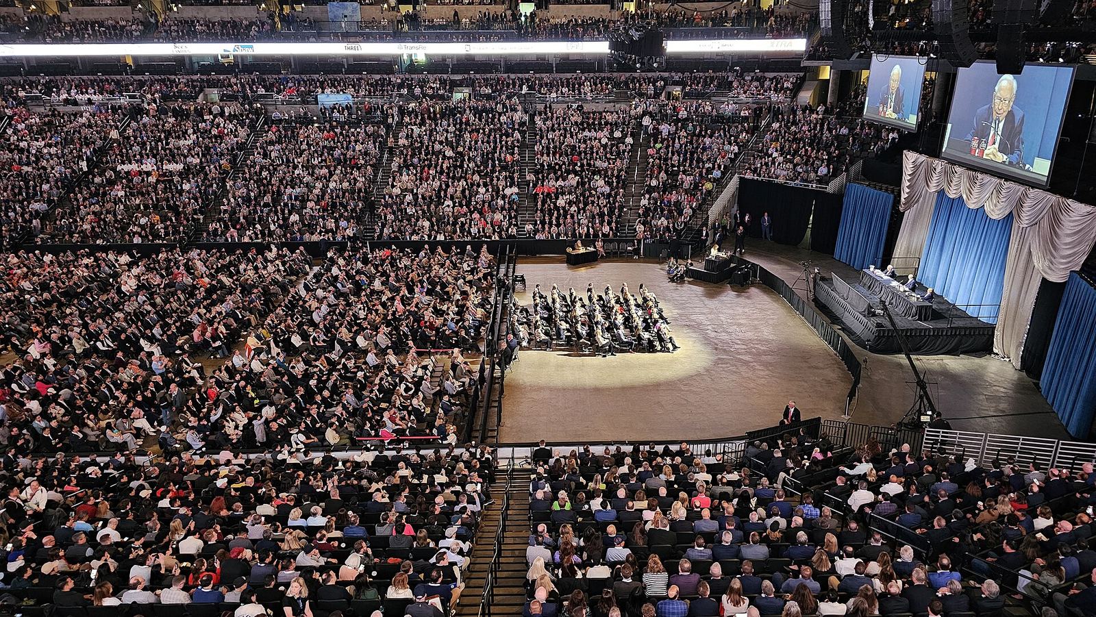 A packed arena at the 2025 Berkshire Hathaway annual meeting in Omaha, with thousands of shareholders in the stands and on the floor, directors seated in rows in front of the stage and Warren Buffett shown on two large screens