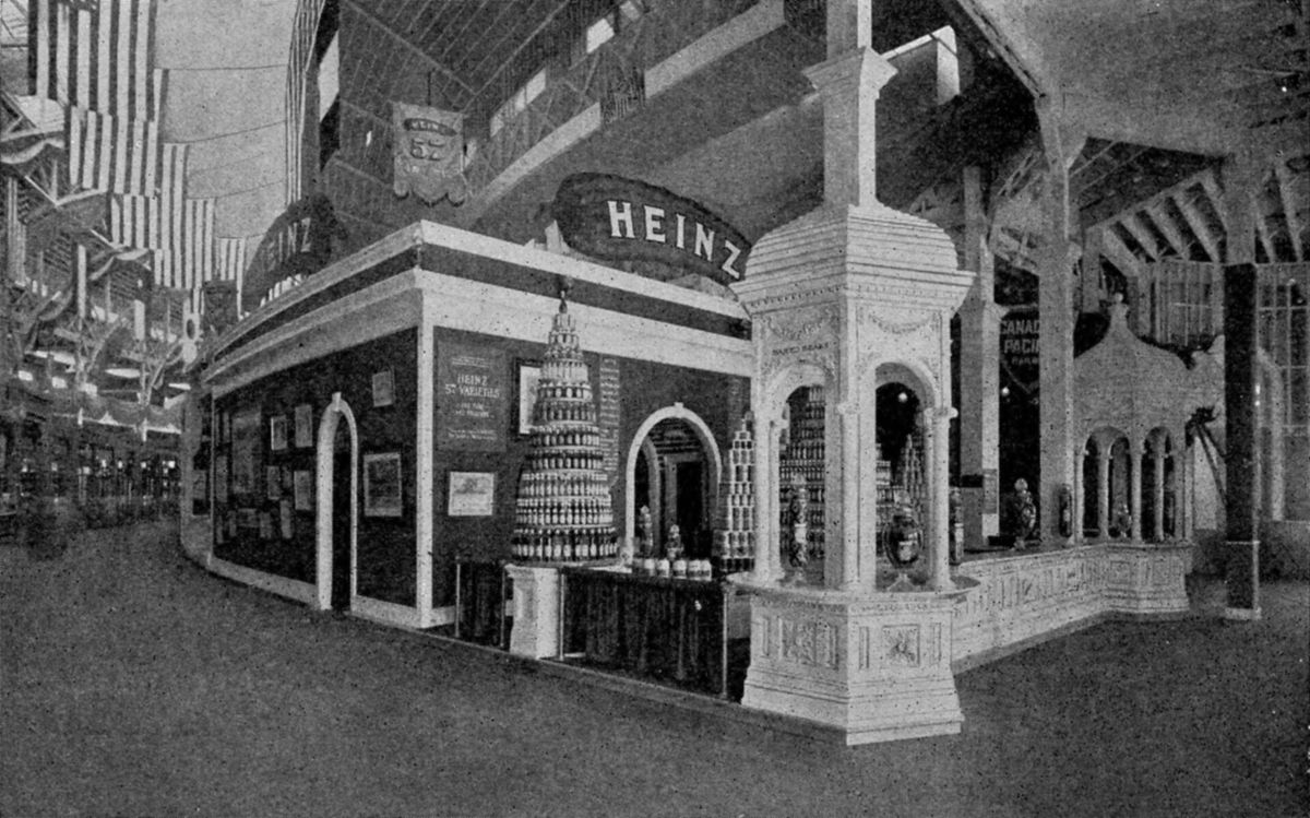 Black and white photograph of a Heinz exhibit booth with a large curved HEINZ sign, a tall pyramid of stacked jars and bottles, an ornate white pillar with arches and display cases, inside an exhibition hall hung with American flags