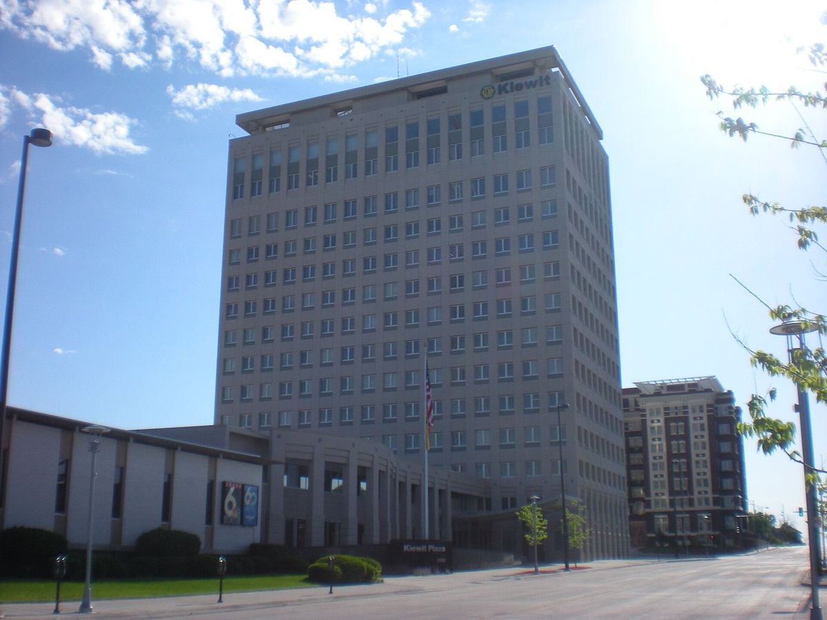 A tall pale office tower with the Kiewit name at the top and a "Kiewit Plaza" sign by the entrance, on a sunny street in Omaha