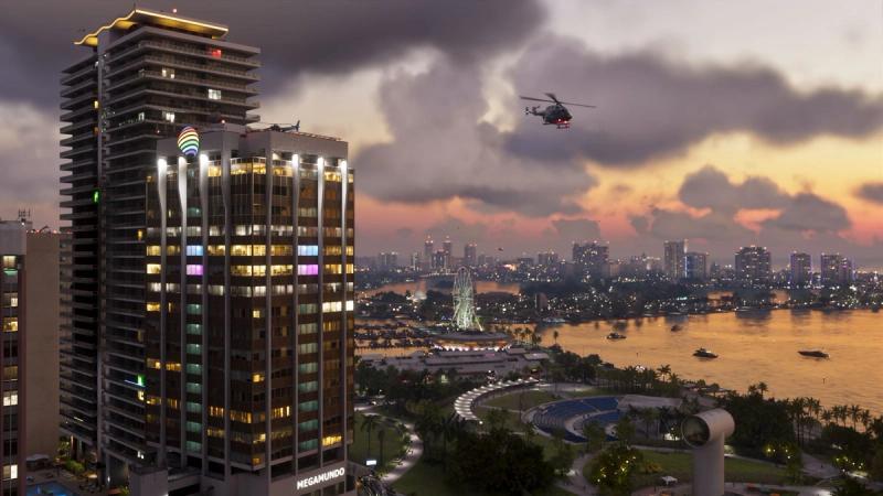 Downtown Vice City at dusk, with a high-rise tower, a waterfront park, a Ferris wheel and a helicopter over the bay — official Grand Theft Auto VI screenshot