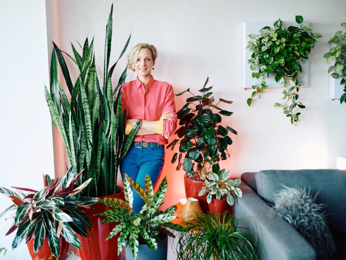 Confident businesswoman in stylish casual attire posing with a collection of indoor plants in a modern office lounge setting