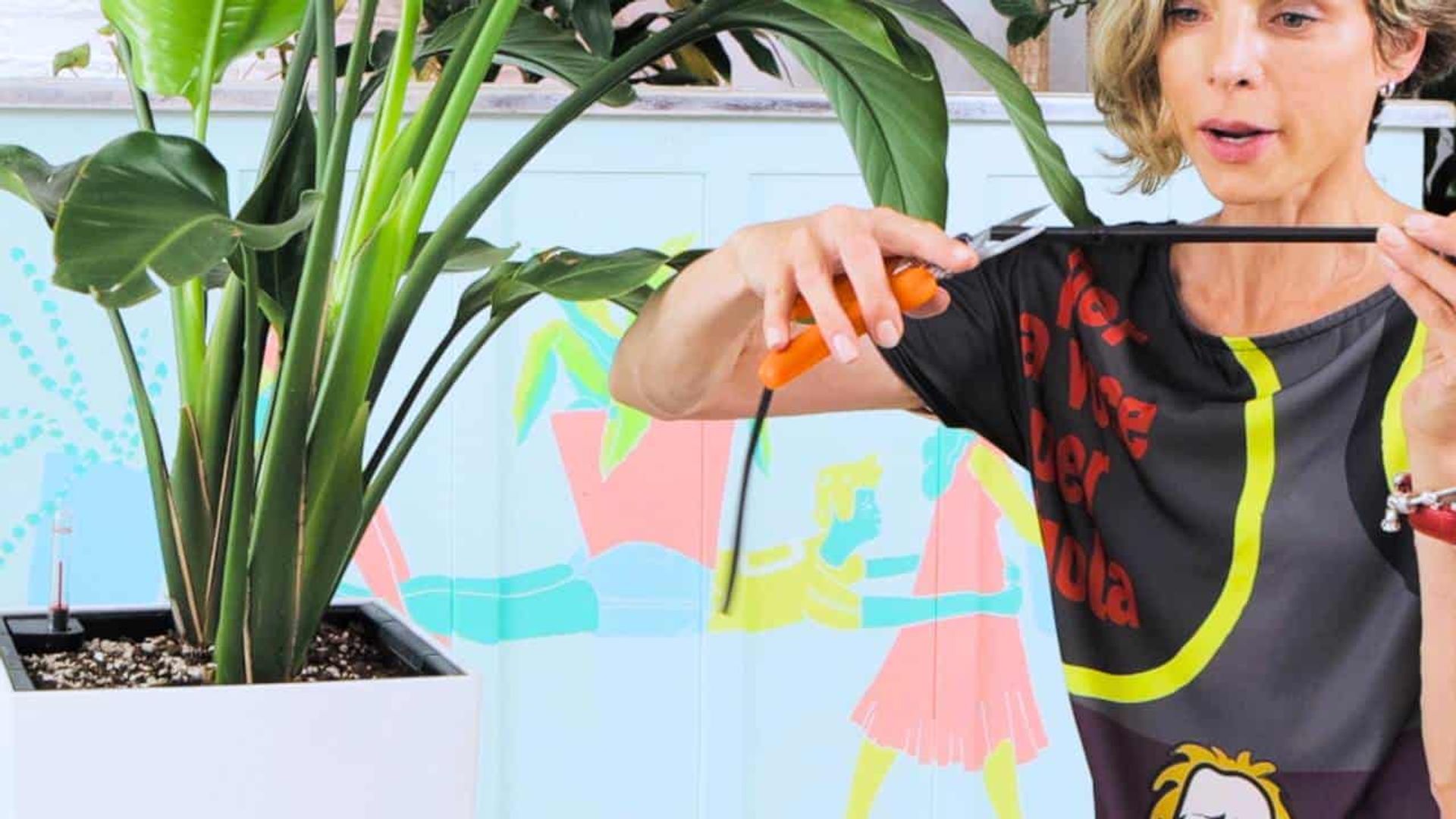 Juliette in a colorful top using orange-handled pruners to cut a bamboo stake, with large green plants and a whimsical mural in the background.