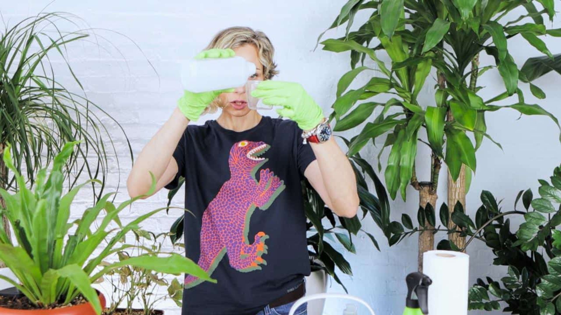 Juliette in a dinosaur t-shirt and green gloves carefully measures a homemade plant pest treatment solution of rubbing alcohol, mild dish soap, and water, against a backdrop of lush indoor plants.