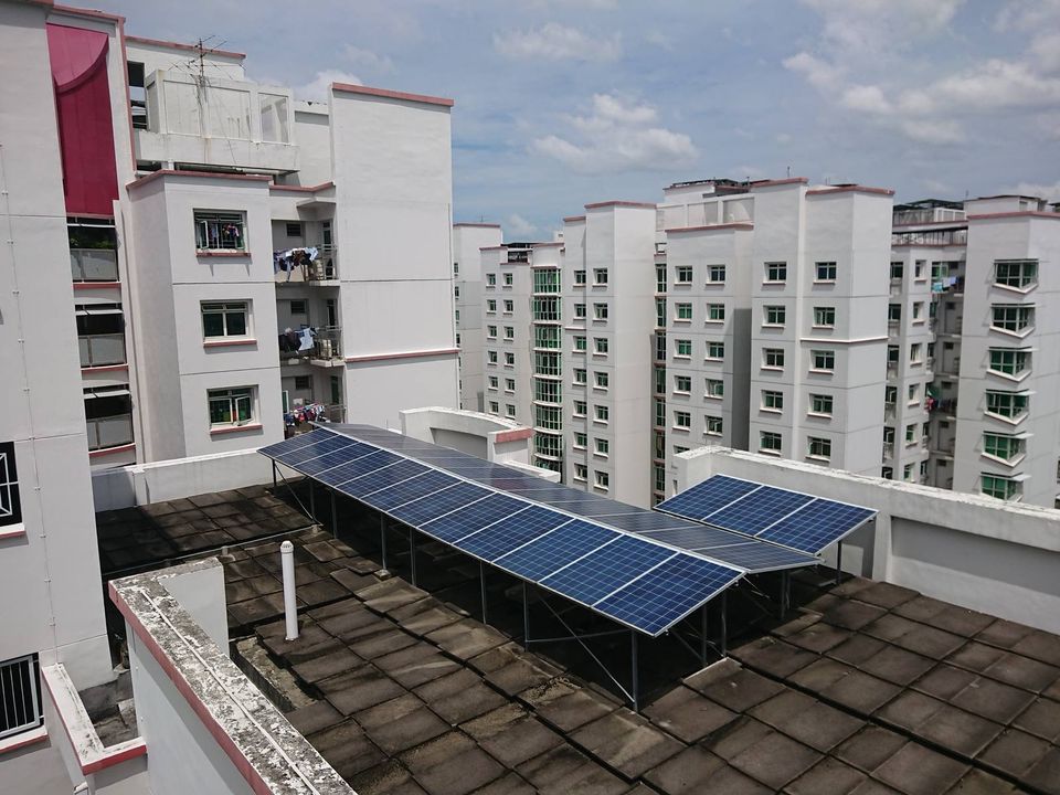 Rows of solar panels mounted on the rooftop of an HDB public housing block in Punggol, Singapore, with other residential blocks behind.