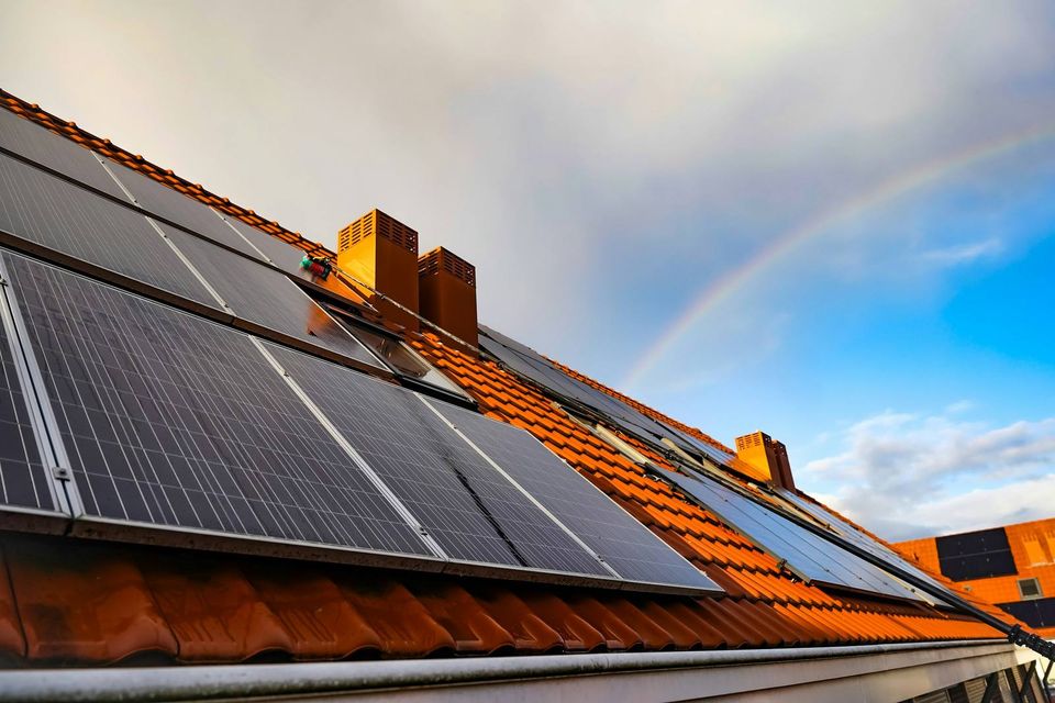 Solar panels fixed to an orange clay-tiled house roof under a cloudy sky with a faint rainbow.