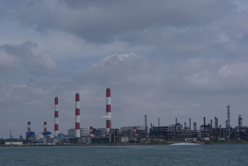 Red-and-white striped chimneys and petrochemical plant structures on Jurong Island seen across the water under a cloudy sky.